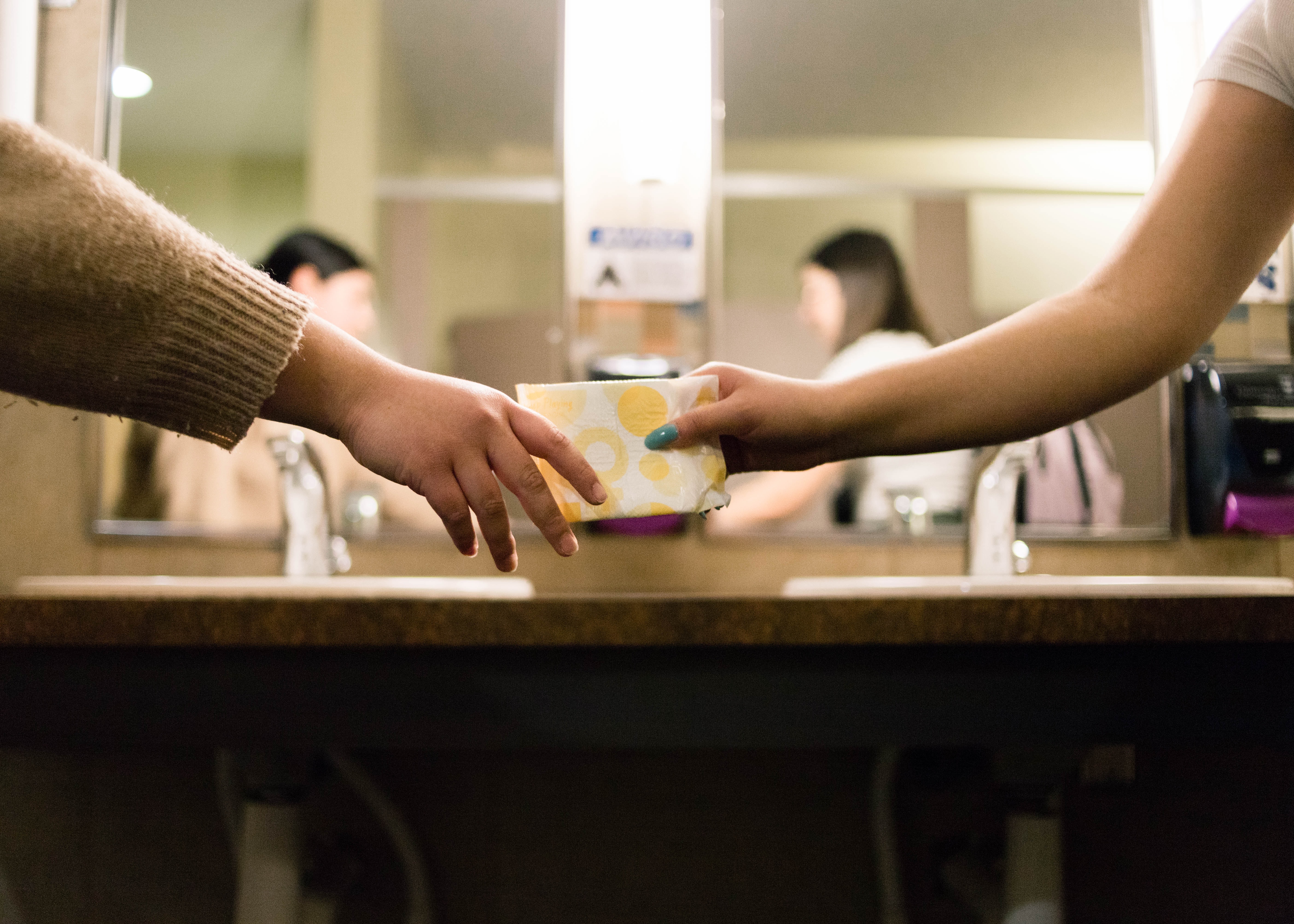 Two people inside a bathroom in front of the bathroom sink. In the middle of the image, one person is passing a pad to the other person.